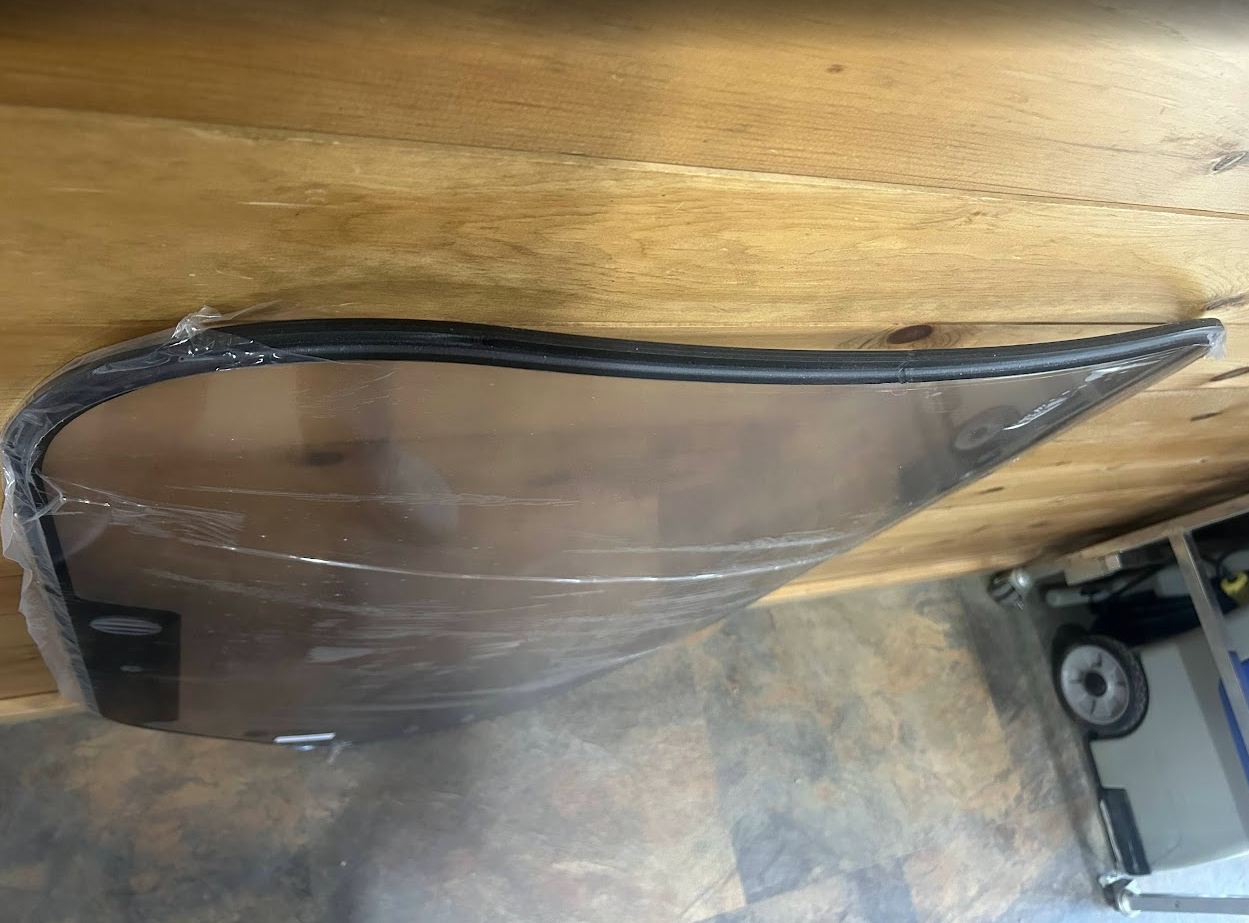 Top edge view of Bobcat M Series 2-hole clear polycarbonate forestry windshield in protective wrap, showing curved top of black door seal frame