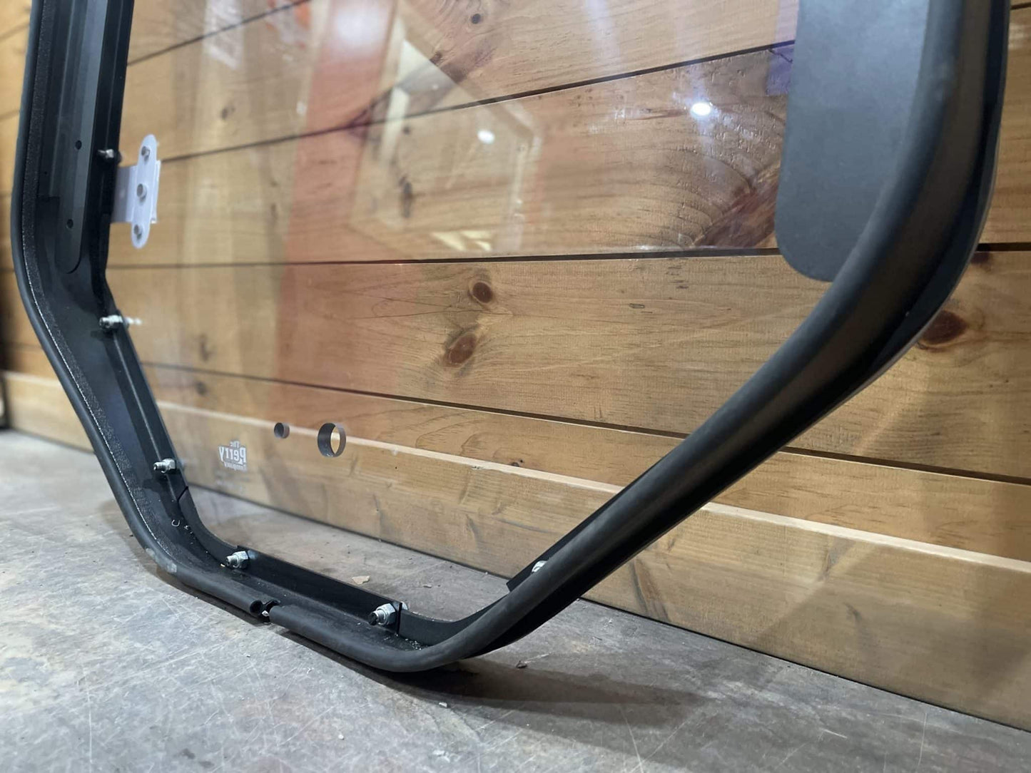 Close-up of Bobcat M Series windshield frame lower corner showing hinge mount bracket, wiper arm mount, and bottom rail detail