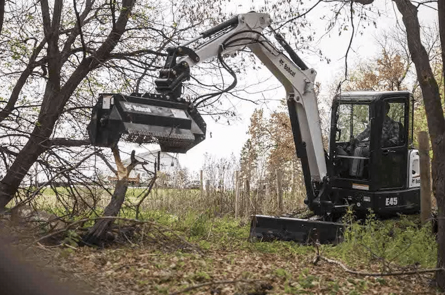 Bobcat E45 mini excavator with forestry mulcher attachment operating in wooded area, showing enclosed cab with polycarbonate windshield