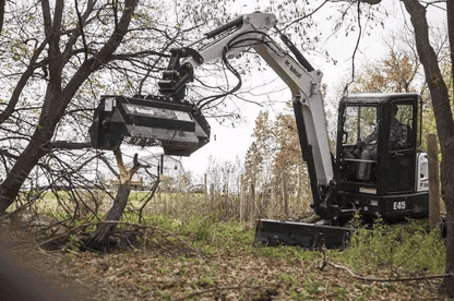 Bobcat E45 mini excavator with forestry mulcher attachment operating in wooded area, showing enclosed cab with polycarbonate windshield