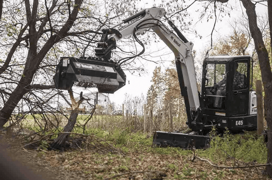 Bobcat E45 mini excavator with forestry mulcher attachment operating in wooded area, showing enclosed cab with polycarbonate windshield