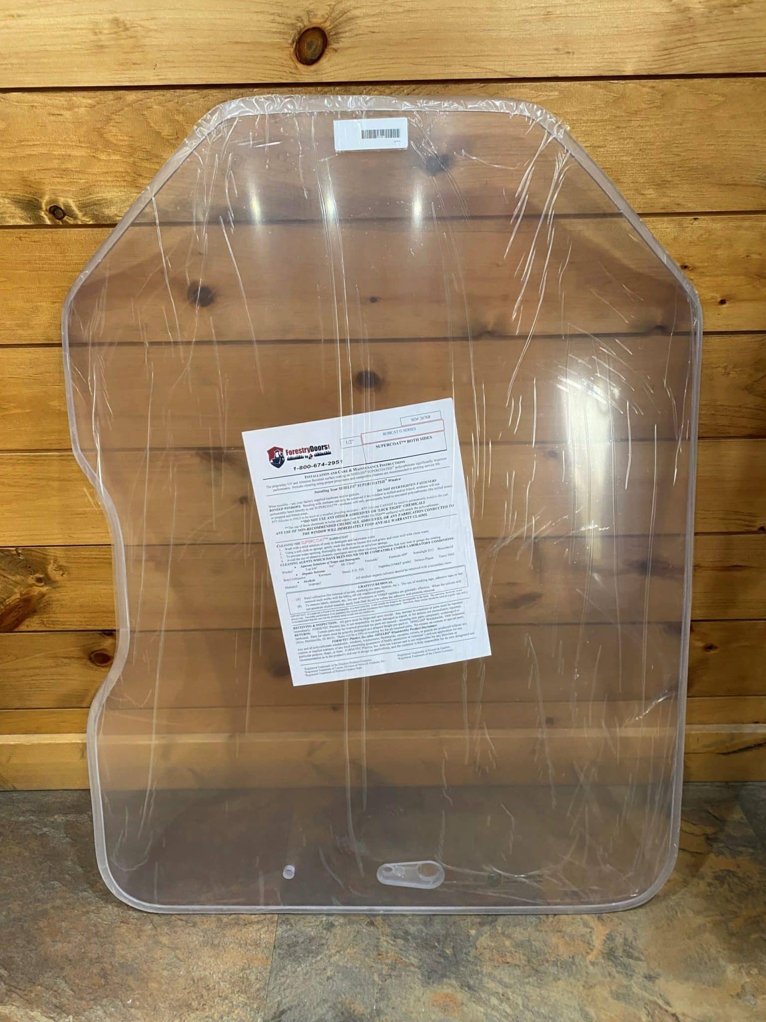 Bobcat G Series clear polycarbonate windshield replacement in protective wrap with installation instruction sheet, leaning against wood wall