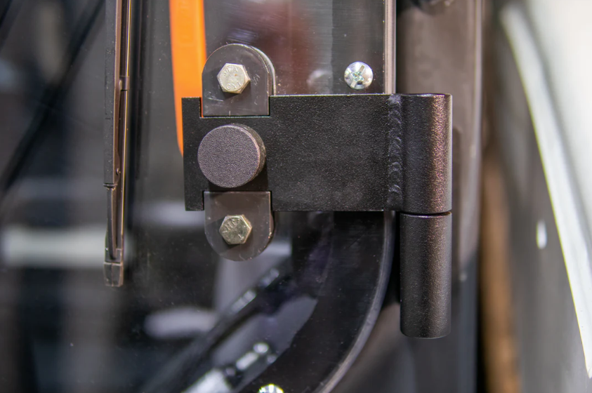 Close-up of Bobcat M Series forestry door hinge assembly showing steel bracket, pivot pin, and mounting bolts