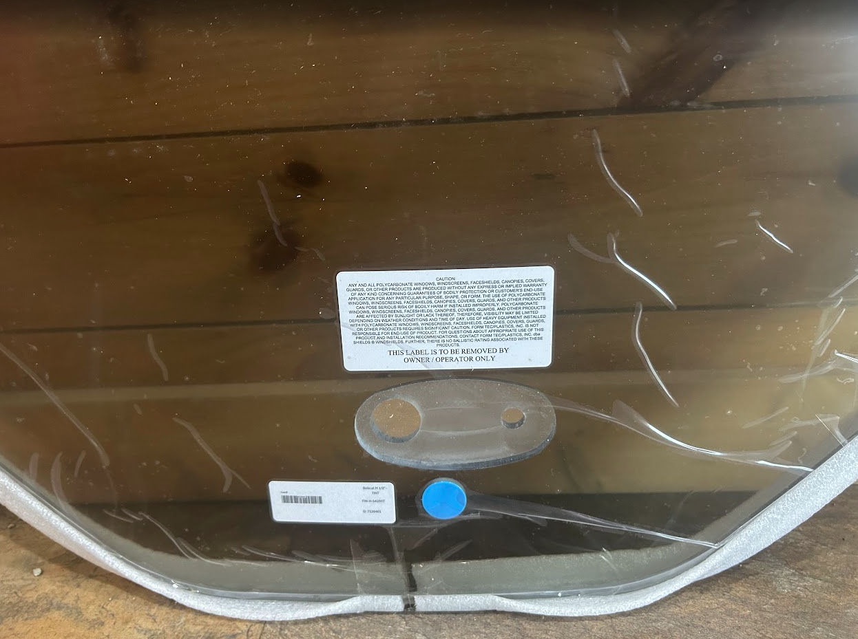 Bobcat M Series tinted windshield interior view showing caution label, wiper mount, wiper fluid cap, and part barcode