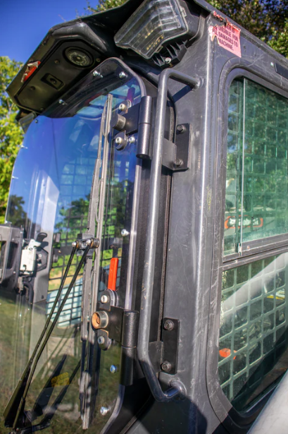 Bobcat R Series skid steer cab side view with door frame and windshield installed, showing wiper arm and hinge hardware