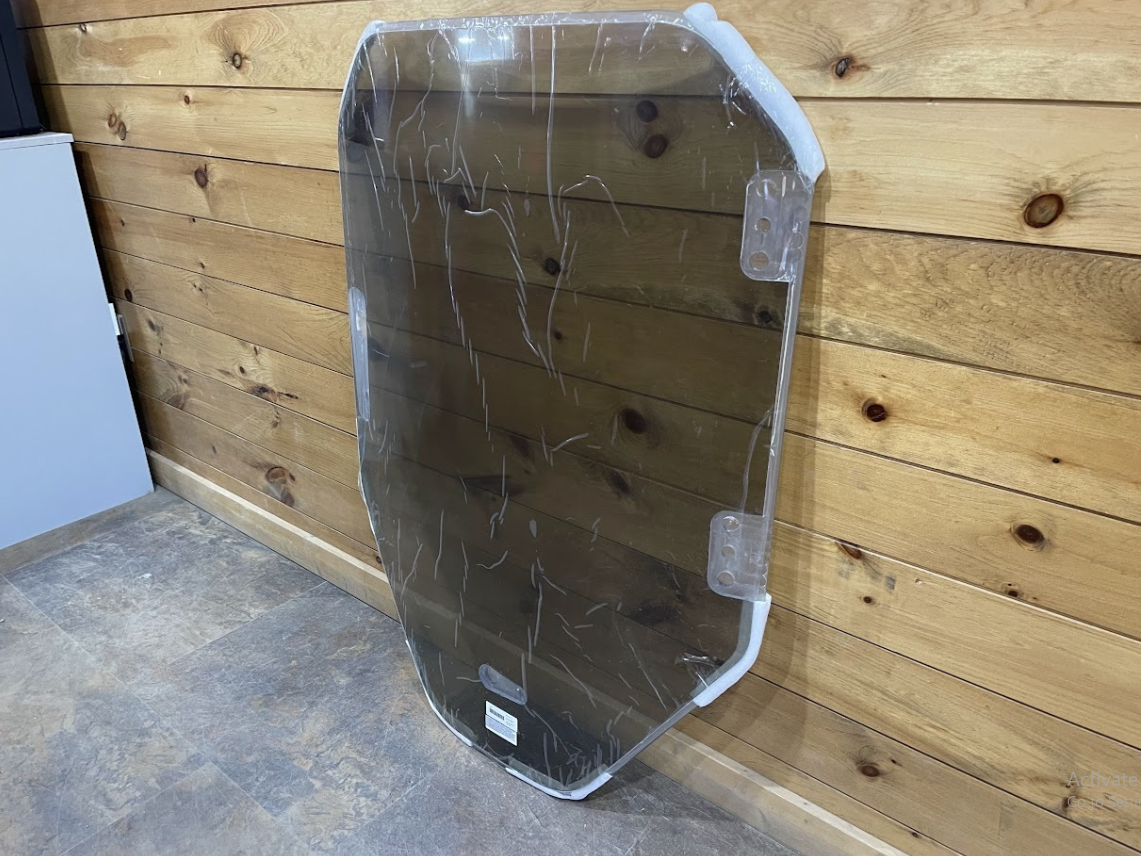 Bobcat R Series polycarbonate windshield leaning against wall, showing side angle and mounting bracket tabs
