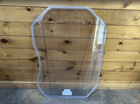 Caterpillar B Series clear polycarbonate windshield replacement panel standing upright against wooden wall with protective wrapping