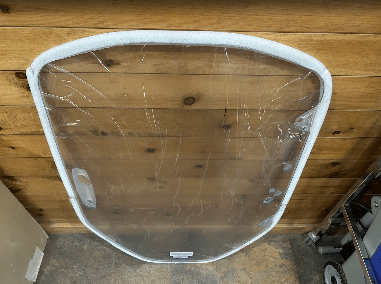 Clear polycarbonate Caterpillar skid steer windshield laid flat on a wood plank floor, shown from a different angle