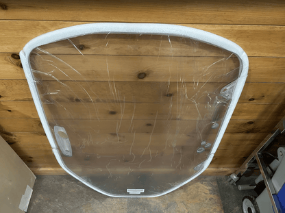 Clear polycarbonate Caterpillar skid steer windshield laid flat on a wood plank floor, shown from a different angle