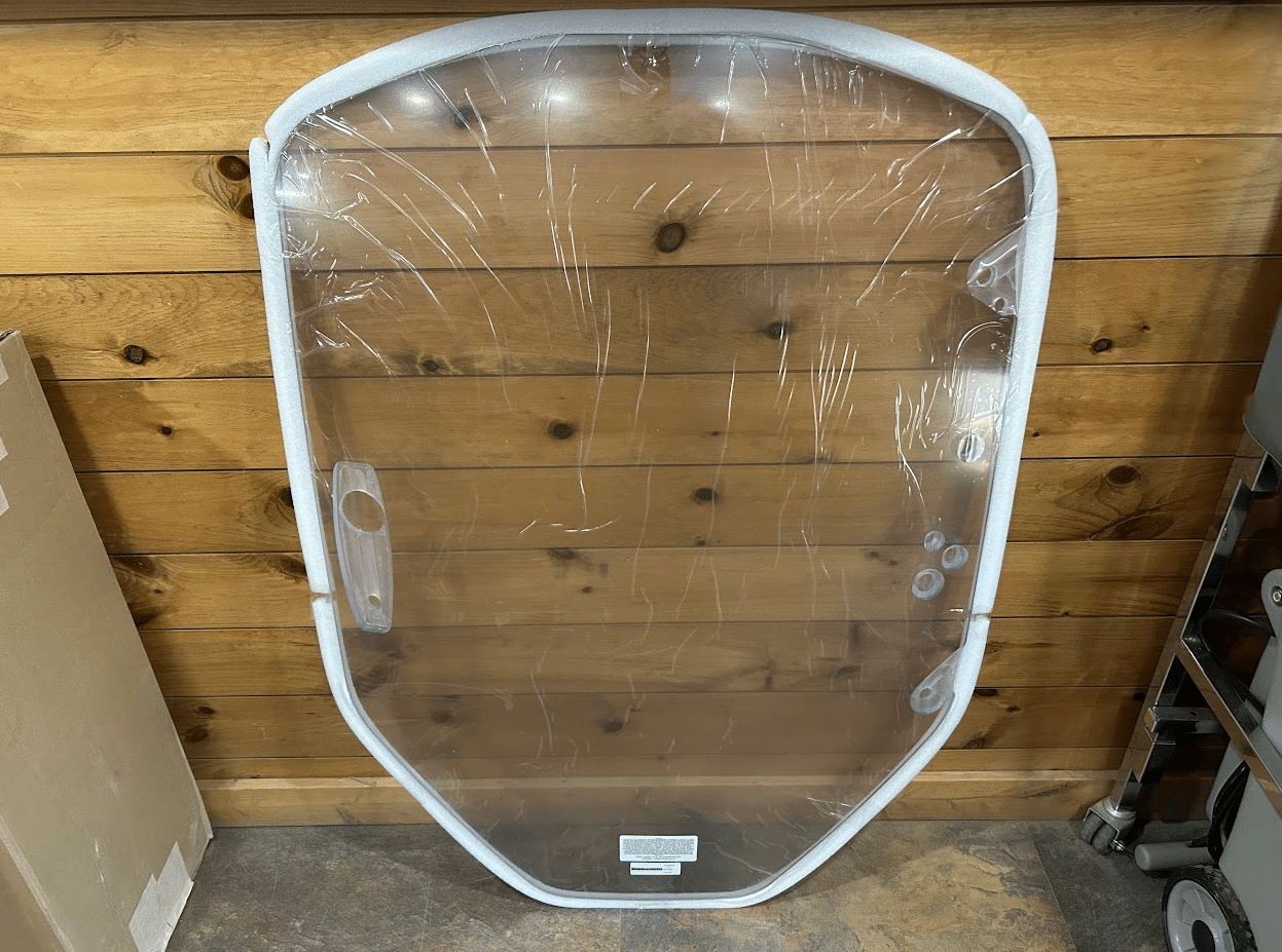 Clear polycarbonate Caterpillar skid steer windshield laid flat on a wood plank floor with protective film still on