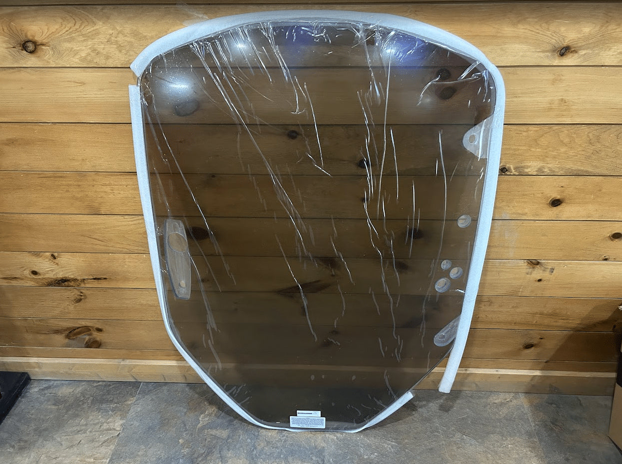 Tinted polycarbonate Caterpillar skid steer windshield leaning against a wood plank wall, shown fully from the front