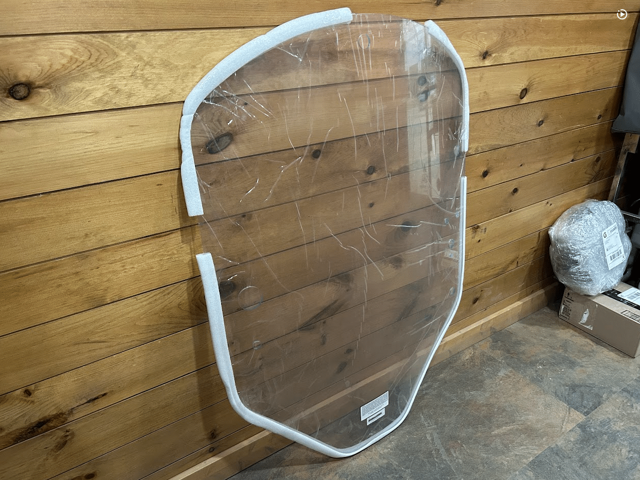 John Deere G Series small frame clear polycarbonate windshield with white frame leaning against wooden wall, side angle view