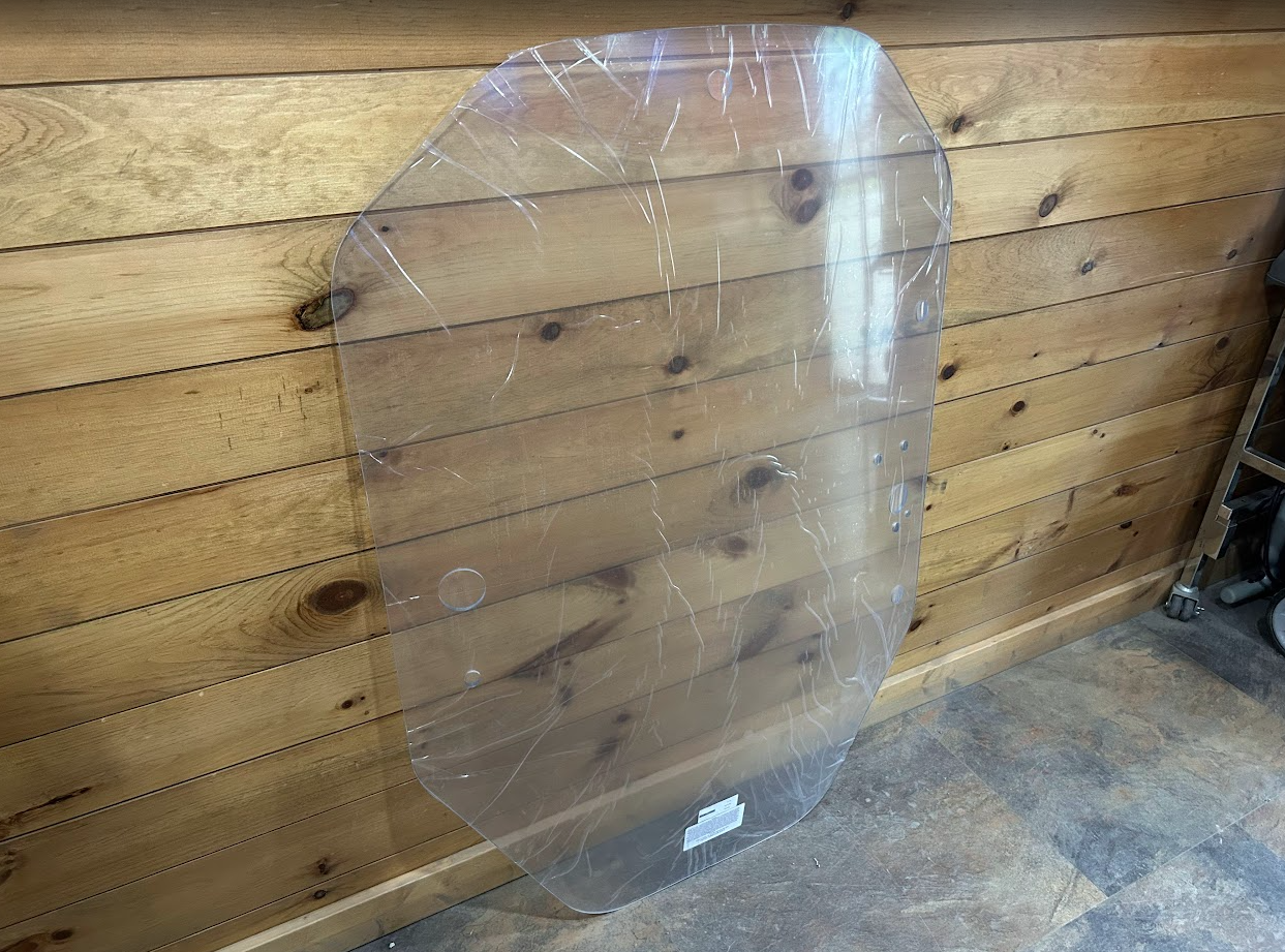 John Deere P Series clear polycarbonate windshield angled right view showing panel profile against wood wall