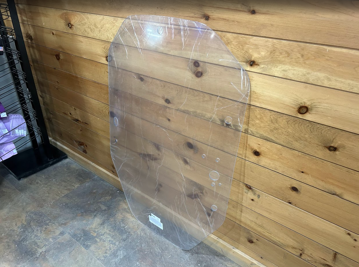 John Deere P Series clear polycarbonate windshield leaning against wood wall in workshop setting, no frame