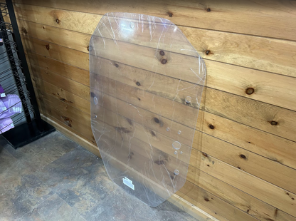 John Deere P Series clear polycarbonate windshield leaning against wood wall in workshop setting, no frame