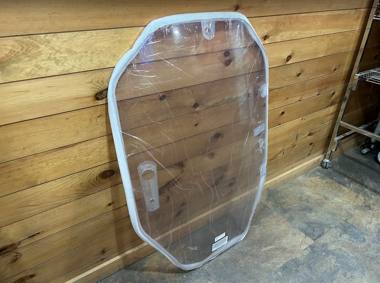 John Deere P Series windshield shown leaning at an angle, plastic wrap on, with hinge hardware visible on left