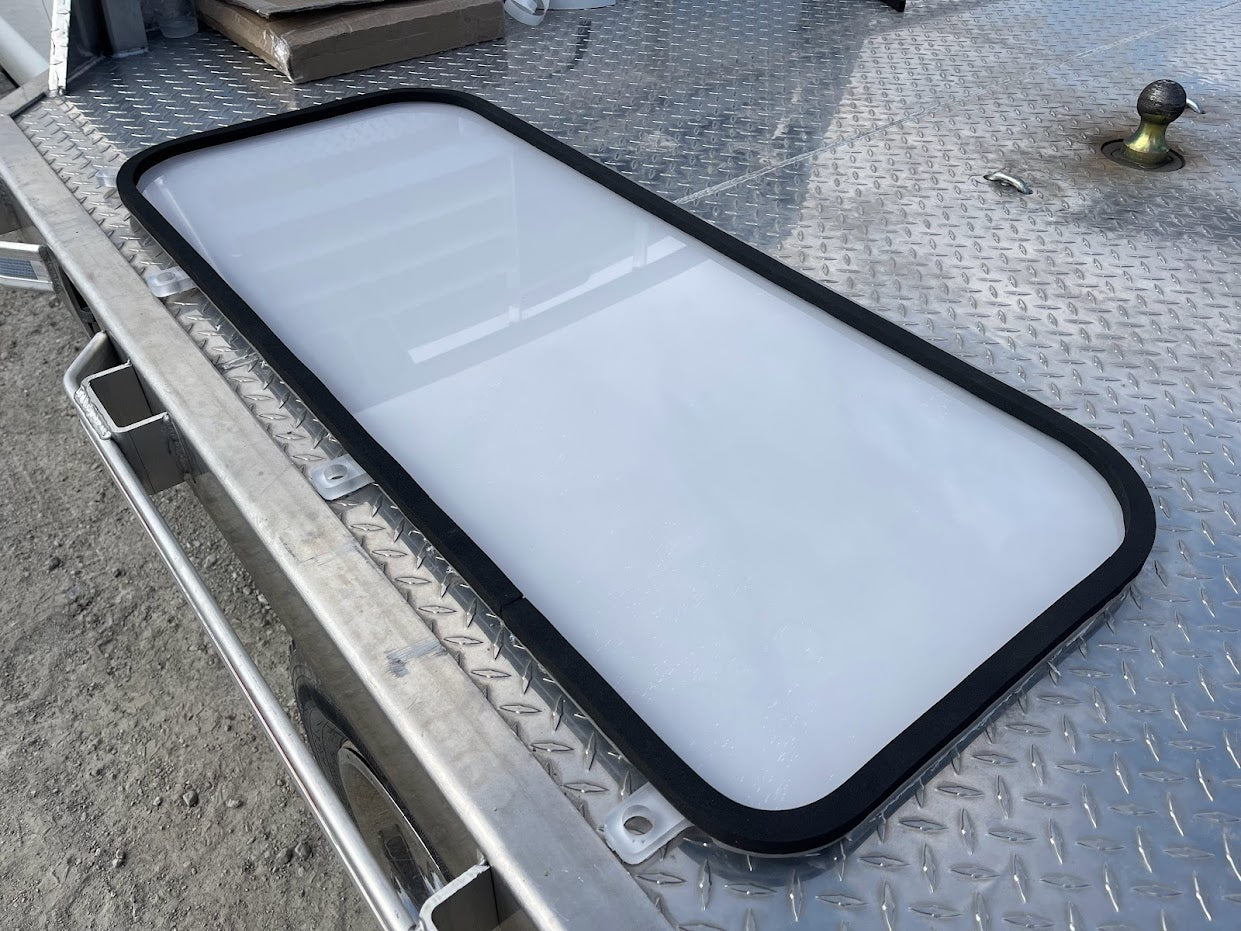 Kubota SVL clear polycarbonate lower side window panel with black rubber seal installed, laid on diamond-plate surface